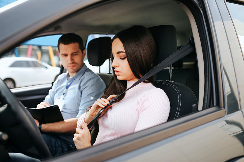 Male auto instructor takes exam in young woman. Brunette hold hands on seat belt and lock it. Young man sit besides with exam papers.
